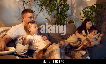 Gemütliche Familie, die sich unter der Decke entspannt, während sie gemeinsam im Wohnzimmer einen Film ansieht. Stockfoto