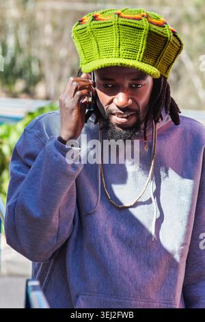 Porträt eines Schwarzen rastafariers mit langen Dreadlocks und einem Spitzbart, der auf einem Smartphone spricht und einen großen grünen Häkelhut mit Rot und du trägt Stockfoto