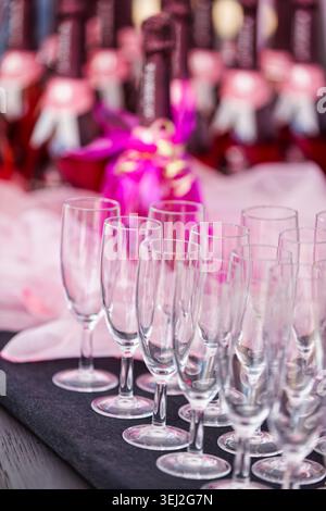 Sektflöten auf einem schwarzen Tisch mit verschwommenen Sektflaschen, die mit rosa Bändern und weichem rosa Stoff verziert sind. Stockfoto