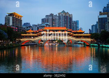 Symmetrischer Blick bei Nacht auf die beleuchtete traditionelle Anshun überdachte Brücke über den Jin Fluss mit Wolkenkratzern Hintergrund, Chengdu, Sichuan, China Stockfoto