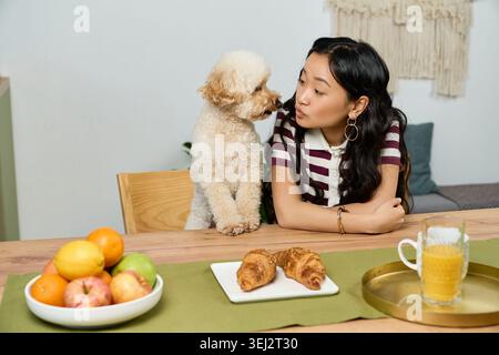 Eine junge Frau interagiert spielerisch mit ihrem Pudel zwischen frischem Obst und Gebäck. Stockfoto