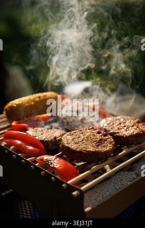 Burger-Patties auf Pflanzenbasis und verschiedene Sommergemüse brutzeln auf einer nachhaltigen Grillbox aus Karton mit verschwommenem grünem Hintergrund. Stockfoto