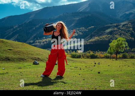 Junge blonde Frau in orangefarbenen Hosen und schwarzem Oberteil, die einen großen Lautsprecher auf der Schulter in einer riesigen Berglandschaft hält Stockfoto