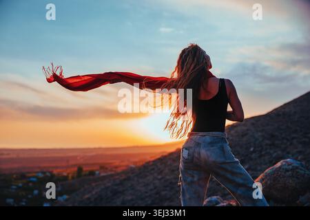 Junge blonde Frau mit langen Haaren und einem fließenden roten Schal, die bei Sonnenuntergang in den Bergen steht Stockfoto