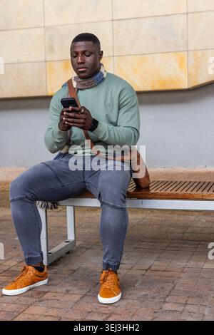 Afroamerikaner sitzt auf einer Bank an einer gefliesten Wand, hält ein Smartphone und trägt orangefarbene Sneakers Stockfoto