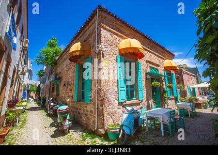 Ayvalik - Türkei, 17. Mai 2026, historisches Steinrestaurant-Gebäude mit türkisfarbenen Rollläden und Motorrollern auf Cunda Island Stockfoto