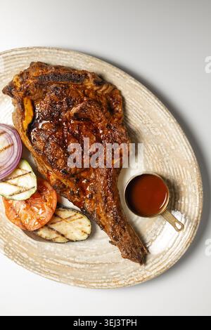 Ein Blick von oben auf ein saftiges gegrilltes Tomahawk-Steak auf dem Knochen, serviert mit Barbecue-Sauce und geröstetem Gemüse auf einer Keramikplatte Stockfoto