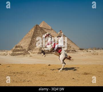 Dynamische Action-Aufnahme eines Mannes, der auf einem dekorierten weißen Pferd reitet und sich dramatisch vor den berühmten Großen Pyramiden von Gizeh in der ägyptischen Wüste aufhält Stockfoto