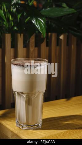 Essen, Ein hohes Glas Eiskaffee steht auf einem Holztisch, gekrönt mit einer Schicht Schokoladenpulver. Üppig grüne Pflanzen bilden eine lebendige Kulisse, Kreation Stockfoto