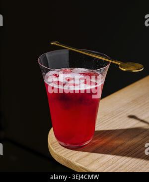 Essen, Ein lebendiges rotes Getränk wird in einem klaren Glas mit Eiswürfeln serviert. Ein goldener Löffel liegt auf dem Rand und sorgt für einen eleganten Look vor dem einfachen Stockfoto