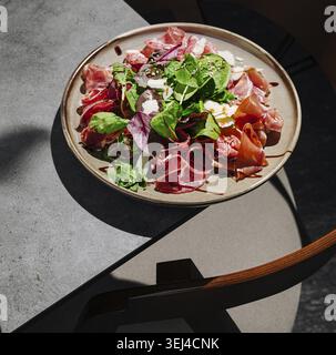 Essen, Ein lebendiger Teller gefüllt mit verschiedenen Wurstsorten und frischem Gemüse auf einem eleganten Tisch. Sonnenlicht wirft weiche Schatten und verbessert so die einladende Pre Stockfoto