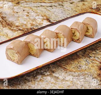 Essen, sechs Bananenbrötchen sind ordentlich auf einer weißen Platte angeordnet. Die Rollen haben eine weiche, braune Außenschicht und sichtbare Bananenscheiben innen. Die Natur Stockfoto