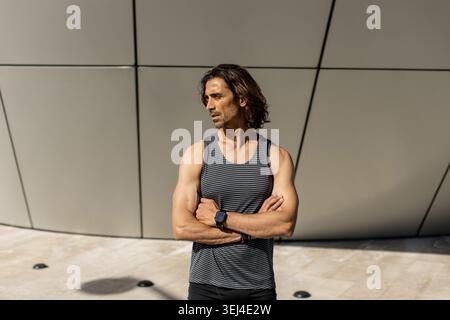 Ein Mann mit langen Haaren steht mit überkreuzten Armen und zeigt lässige Mode, fast schlankes, geometrisches Gebäudedesign unter hellem Sonnenlicht. Stockfoto