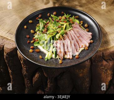 Essen, Ein frischer Salat mit geschnittenem Fleisch, Blattgemüse und getrockneten Früchten steht auf einem schwarzen Teller. Das Gericht ist wunderschön auf einem rustikalen hölzernen t angeordnet Stockfoto