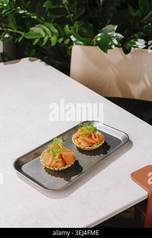 Essen, zwei feine Meeresfrüchte-Tartlets sind elegant auf einem glänzenden Tablett angeordnet. Das helle Grün der Pflanzen im Hintergrund bereichert das moderne Essen im Stockfoto