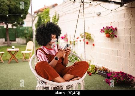 Frau, die sich im Hängesessel entspannt und im Garten im Garten des Hinterhofs ihr Smartphone durchsucht Stockfoto