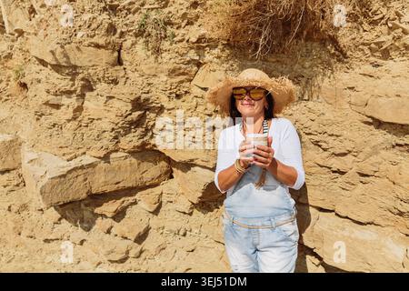 Reife Frau mit Strohhut, lächelt mit Kaffee an den Felsen der Küste Stockfoto