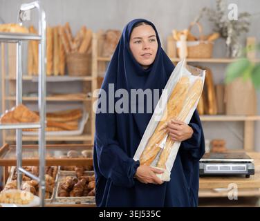 Porträt eines orientalischen Mädchens in Abaya mit frischen Baguettes in der Hand in der Bäckerei Stockfoto