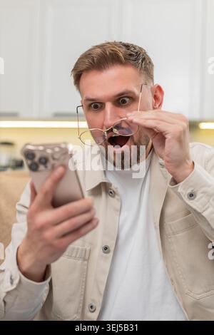 Ein junger Mann zu Hause liest mit einer Brille winzige Texte auf dem Smartphone und staunt über die Geschichte und Neuigkeiten. Der kaukasische Kerl auf dem Sofa sieht starke Aufmerksamkeit im Boten, zeigt Freude Erleichterung und helle Wow-Überraschung auf einmal Stockfoto