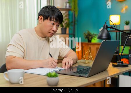 Asiatischer Mann, der zu Hause am Tisch sitzt, nimmt an einem Online-Kurs auf Laptop Teil, schreibt Notizen und erklärt der Kamera, wie sie sich zeigt. Der Chinese spricht selbstbewusst, mit ausdrucksstarken Gesten, um ansprechende Online-Lektion zu erstellen. Stockfoto