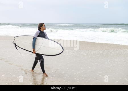 Junge Surferin im Taucheranzug, Surfbrett unter dem Arm, am Strand nach dem morgendlichen Surfen Stockfoto