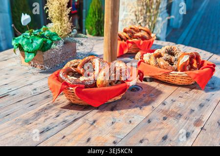 Körbe gefüllt mit frisch gebackenen Brezeln auf einem rustikalen Holztisch im Freien. Brezeln sind goldbraun, mit Salz und Samen bestreut und mit Beilagen Stockfoto