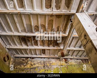 Im verlassenen Innenraum ist eine Decke mit abblätternder Farbe und Sichtbeton in einem verlassenen Lagerhaus zu sehen. Hängende Metallbefestigungen tragen zum verfallenen Aussehen bei Stockfoto