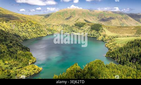 Blick aus der Vogelperspektive auf einen atemberaubenden türkisfarbenen See inmitten üppiger grüner Hügel. Das Wasser reflektiert den Himmel, umgeben von dichter Vegetation und rollendem Berg Stockfoto