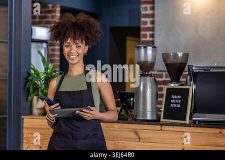 Afroamerikanerin in ihren Zwanzigern mit schwarzer Schürze, die Tablet und Telefon mit Espresso hält Stockfoto