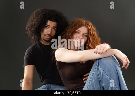 Junges, vielfältiges Paar, das auf grauem Hintergrund sitzt und Verbindung und Glück annimmt Stockfoto