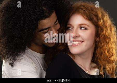 Lächelnder junger Mann, der eine Frau mit lockigen roten Haaren umschließt und sich beide glücklich ansieht Stockfoto