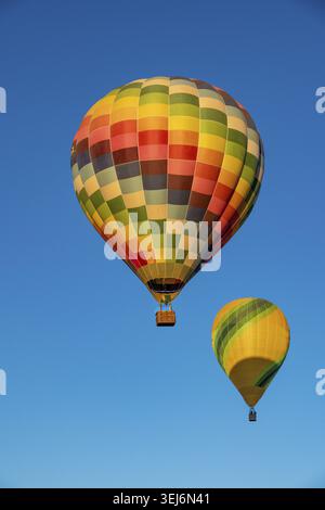 Zwei bunte Heißluftballons steigen in den klaren blauen Himmel. Das eine hat ein lebhaftes Karomuster, das andere ist gelb-grün gestreift Stockfoto