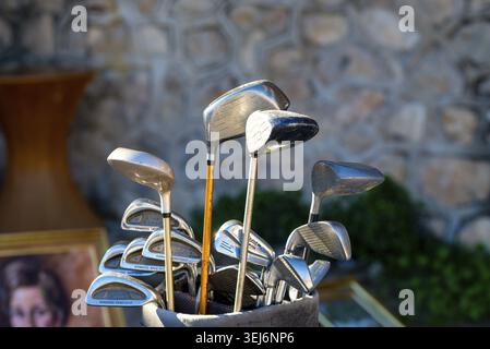 Xabia - Valencia Community - Spanien - 22.11.2025: Golfschläger und Bügeleisen stehen in einer Tasche in der Nähe, beleuchtet von niedriger Sonne, während eine verschwommene Steinmauer und Grün Stockfoto