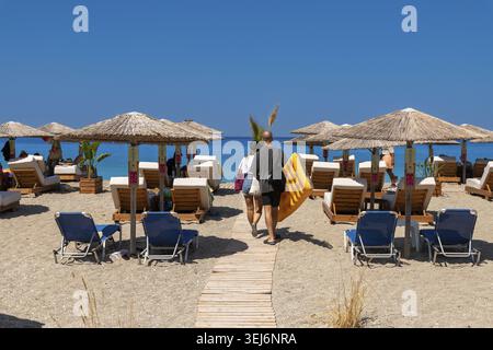 Nydri - Ionische Inseln - Griechenland - 08-09-2025: Zwei Personen gehen auf einem Holzweg am Sandstrand entlang, vorbei an Strohschirmen und -Liegen, mit Yello Stockfoto