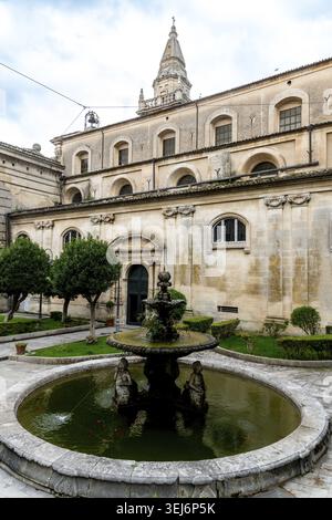 Ein detaillierter Blick auf die Fassade der Kirche San Giovanni Battista mit einem Steinbrunnen im Vordergrund mit Zierbäumen an der Seite Stockfoto