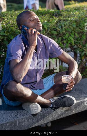 Entspannter junger Schwarzer Mann, der sich mit dem Smartphone unterhalten kann, während er draußen bei einem kalten Getränk am sonnigen Sommernachmittag in authentischer urbaner Lifestyle-Szene sitzt. Stockfoto