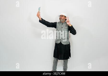 Ein indischer Mann mittleren Alters macht ein Selfie, während er traditionelle muslimische Kleidung trägt, darunter eine graue und schwarze Weste, eine dunkle Hose und eine weiße Mütze. Stockfoto