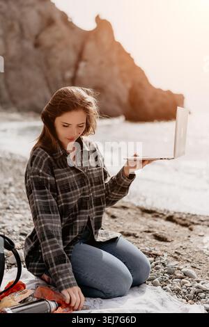 Frau Laptop Strand junge Freelancerin in kariertem Hemd arbeitet fernab an steinigen Küsten mit Klippen während einer Urlaubsreise Stockfoto