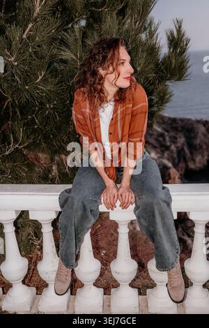Frau Küstenbalustrade junge Frau mit lockigen Haaren, die auf weißem Steingeländer sitzt und zur goldenen Stunde die Klippen überblickt Stockfoto
