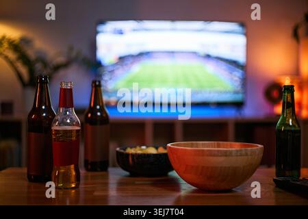Bierflaschen und Snackschalen stehen auf dem Tisch, während Freunde Fußballspiele im Fernsehen verfolgen, die männliche Freundschaft und Freizeit repräsentieren. Ideal für Sportbeobachtungen, Bonding und Home Entertainment Stockfoto