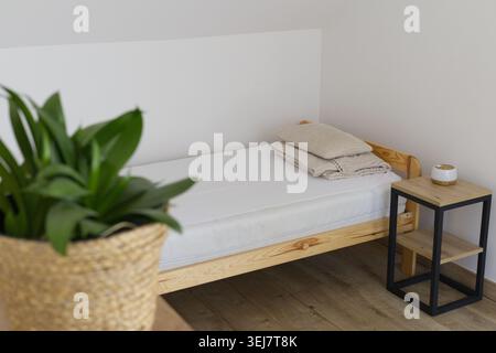 Helles Schlafzimmer im Dachgeschoss mit minimalistischem Holzbett, weißer Bettwäsche, Nachttisch im Loft-Stil und verschwommener, grüner Pflanze im Vordergrund Stockfoto