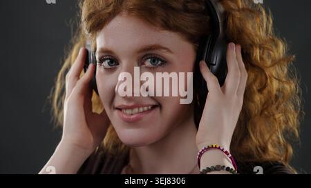 Eine junge Frau mit lockigen roten Haaren hört Musik auf den Kopfhörern und lächelt Stockfoto