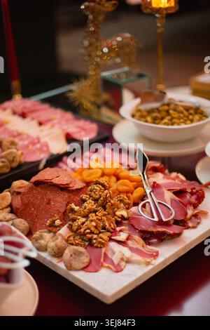 Wurstplatte, beladen mit Salami, Wurstwaren, Walnüssen, getrockneten Feigen und Aprikosen, serviert mit Zange an einem eleganten Buffet. Stockfoto