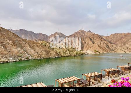 Malerischer Blick auf das Erholungsgebiet Al Rafisah Dam in Sharjah, VAE, mit entspannenden Pavillons am See, grünem Wasser und zerklüfteten Hajar Mountains unter Clou Stockfoto