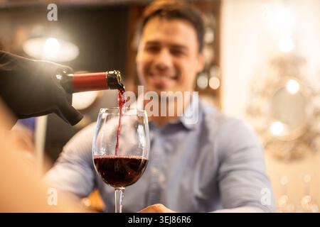 Nahaufnahme eines Rotweins, der während eines romantischen Abendessens in einem gemütlichen Restaurant in ein Glas gegossen wird. Junger Mann lächelt im Hintergrund, während er ein El genießt Stockfoto