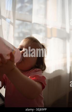 Nahaufnahme eines Kleinkindes, das aus einer rosa Flasche trinkt, in sanfter Morgenbeleuchtung durch transparente Vorhänge, eine warme, gemütliche Hausszene mit sanften Schatten Stockfoto