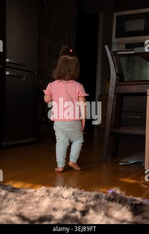 Rückansicht eines Kleinkindes in rosa Hemd und leichten Leggings, der barfuß über warmen Küchenboden aus Hartholz zu einem dunklen Eingang geht, weiches natürliches Licht Stockfoto