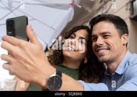 Glückliches junges Paar, das gemeinsam Drinks genießt und ein Selfie mit einem Smartphone auf einer Terrasse im Freien macht. Lässiger Lifestyle-Moment mit Bier, Stockfoto