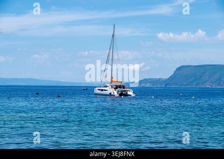 Katamaran vor der Küste von Scilla, Kalabrien, mit verstreuten Bojen und einer entfernten grünen Berglandschaft unter einem teilweise bewölkten Himmel. Stockfoto