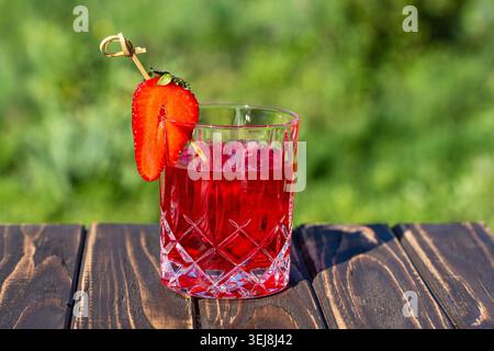Kaltes Getränk aus roten Beeren garniert mit frischer Erdbeere auf einem Holztisch. Helle Sommer-Cocktail-Fotografie mit verschwommenem grünem Gartenhintergrund und na Stockfoto